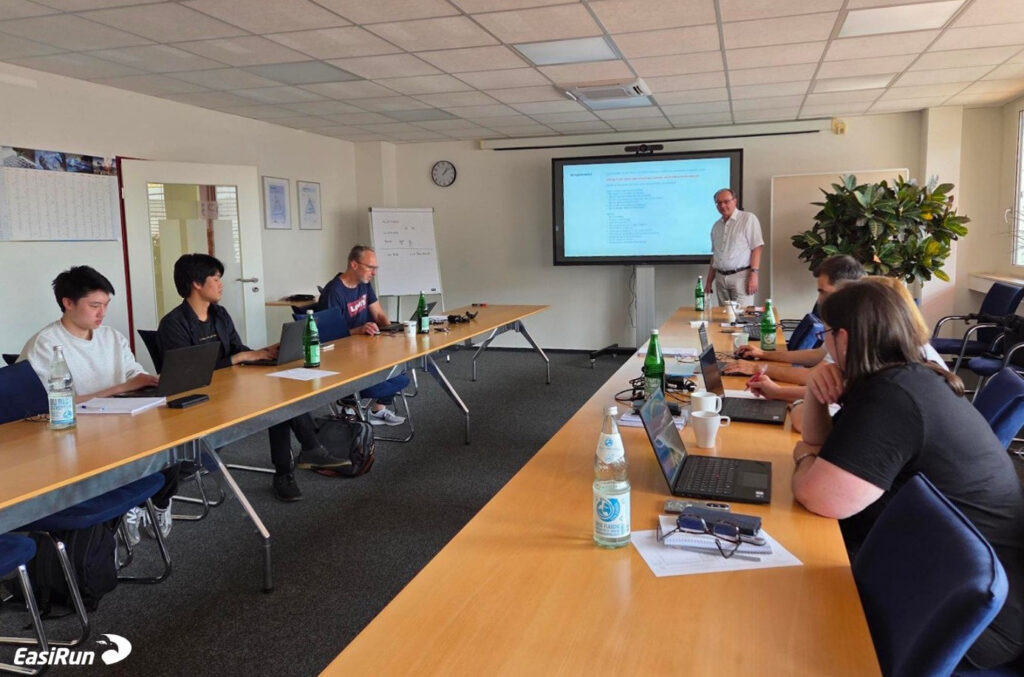 Team meeting in a conference room with a presenter at the front near a projected slide screen and attendees using laptops around a long table.